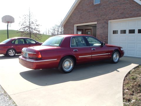 1999 Mercury Grand Marquis - Two red sedans are parked in a driveway in front of a brick house with a white garage door.