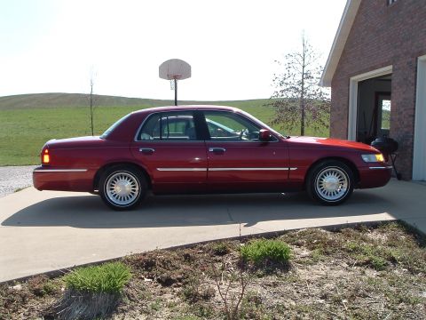1999 Mercury Grand Marquis - A red 1999 Mercury Grand Marquis sedan is parked on a concrete driveway next to a brick garage.