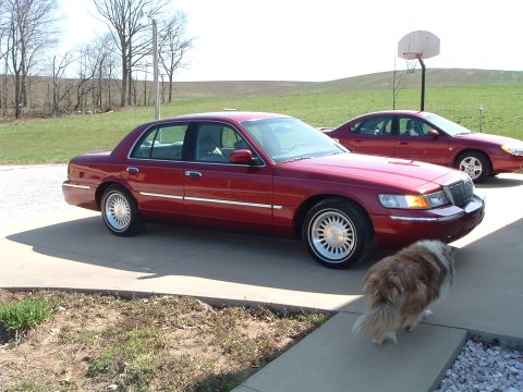 1999 Mercury Grand Marquis - A red 1999 Mercury Grand Marquis is parked on a driveway next to another car, with a dog in the foreground.