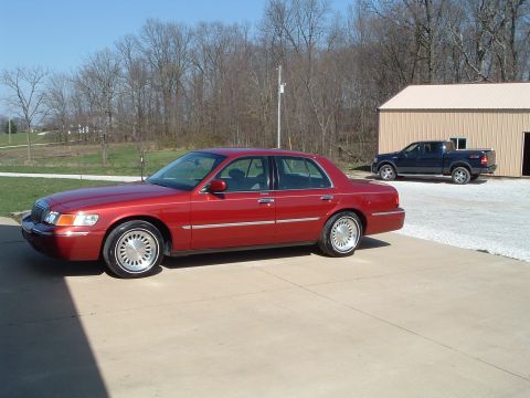 1999 Mercury Grand Marquis - A red 1999 Mercury Grand Marquis sedan is parked in front of a building, with a pickup truck in the background.