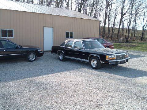 1999 Mercury Grand Marquis - A black 1999 Mercury Grand Marquis is parked next to another sedan in front of a tan building.