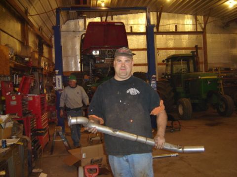 1992 Ford Bronco - A man holds a large exhaust pipe while standing in a garage with a tractor and another vehicle on a lift.