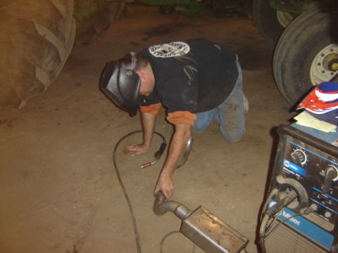 1992 Ford Bronco - A person is welding an exhaust pipe while kneeling on the ground, with parts of a large vehicle and equipment visible.