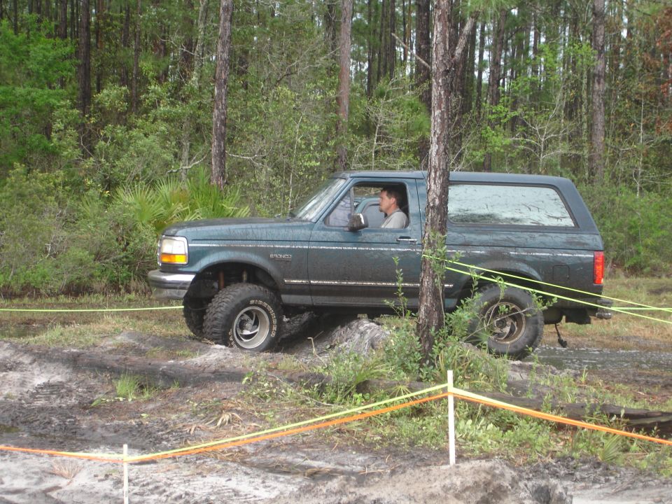 Green Ford Bronco with lifted suspension driving through muddy terrain with trees in background