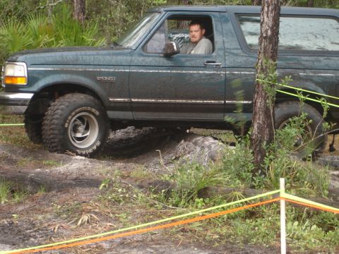1989 Ford Bronco - A man drives a green Ford Bronco through an off-road course with logs and foliage.