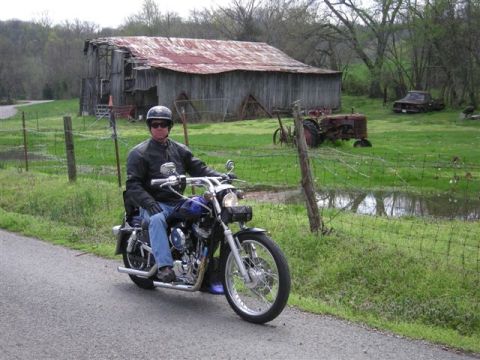 2005 Ford Crown Victoria - A rider on a blue motorcycle travels down a paved road next to a grassy field and an old barn.