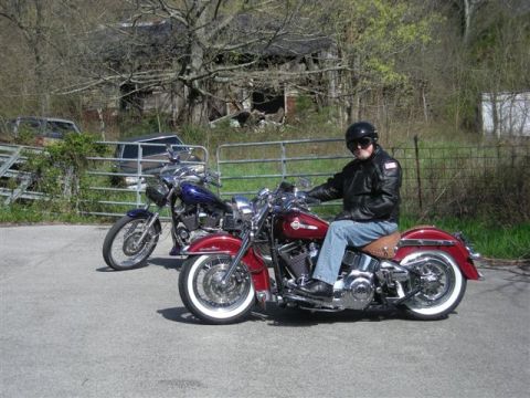 2005 Ford Crown Victoria - Two motorcycles are parked in front of a dilapidated building with trees and a fence.