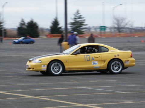 2001 Mercury Grand Marquis - A yellow 1997 Ford Mustang GT coupe with racing decals is seen on a track, with another car in the background.