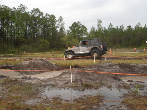 1989 Ford Bronco - A Jeep Wrangler navigates a muddy obstacle course under an overcast sky.