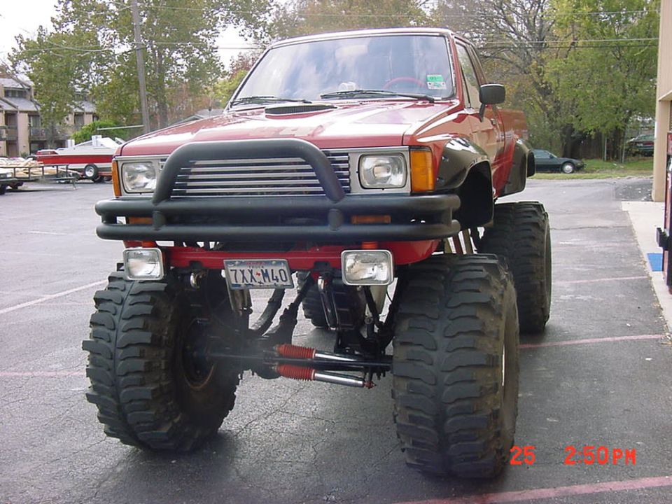 Lifted red pickup truck with large off-road tires and a brush guard. License plate reads 7XX-M40
