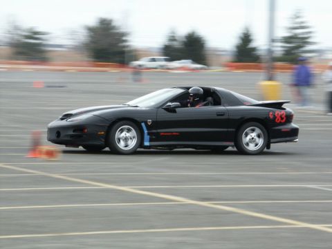 2001 Mercury Grand Marquis - Black Pontiac Firebird with racing number 83 on the rear fender drives on a paved track with orange cones.