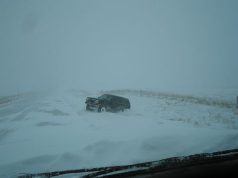 1990 Ford Bronco - A black 1990 Ford Bronco struggles through deep snow on a road during a blizzard, with visibility severely limited.