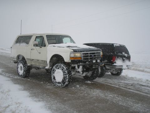 1990 Ford Bronco - Two SUVs, including a white 1990 Ford Bronco, navigate a snow-covered road.
