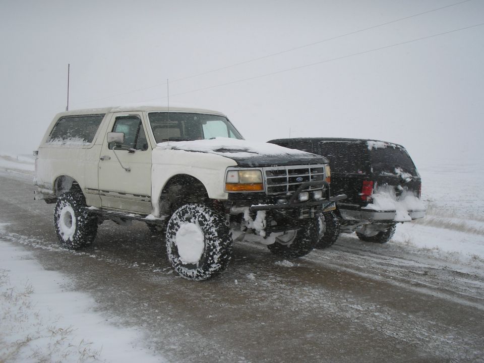 White 1990 Ford Bronco and black SUV driving on a snowy road