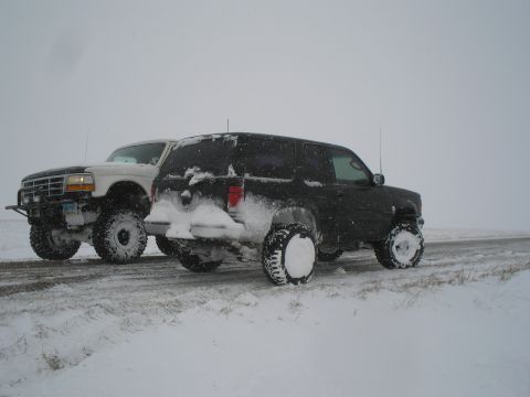 1990 Ford Bronco - Two lifted SUVs are parked on the side of a road during a snowstorm.