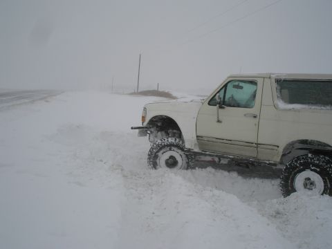 1990 Ford Bronco - A white 1990 Ford Bronco is partially buried in snow, struggling to move on a desolate road during a snowstorm.