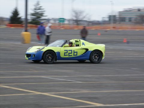 2001 Mercury Grand Marquis - A two-tone yellow and blue Lotus Elise with racing numbers navigates a track, with spectators blurred in the background.