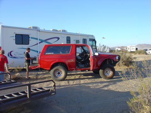 1984 Ford Bronco - A red off-road SUV is parked next to a large RV in a dry, open area with sparse vegetation.