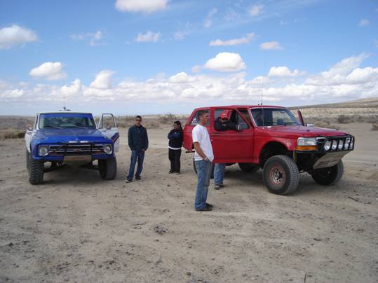 Blue vintage Ford pickup and red Ford prerunner in the desert with people