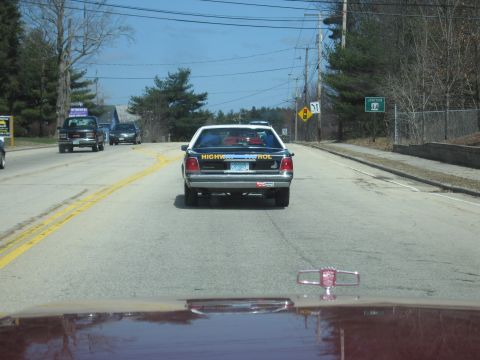1991 Ford Crown Victoria - A highway patrol vehicle is seen from behind on a road, with other cars and trees visible in the background.