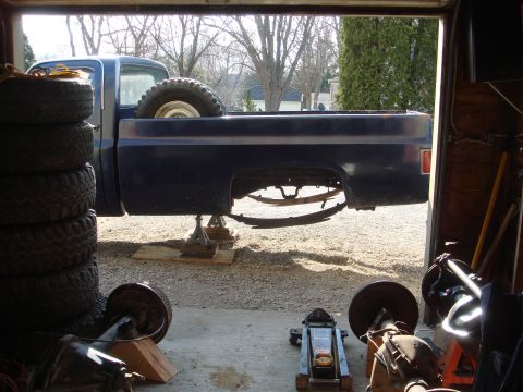 1978 Chevrolet C/K Pickup - A blue pickup truck is raised on jack stands in a garage, with spare tires and automotive parts visible.