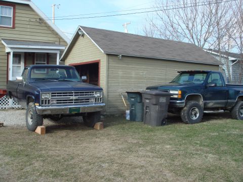 1978 Chevrolet C/K Pickup - Two pickup trucks are parked in a yard next to a garage and a house. The blue truck is on blocks.