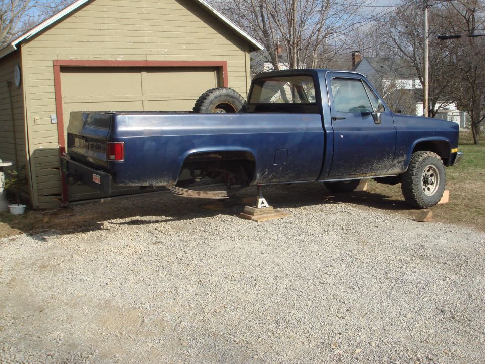 Side view of a blue 1978 Chevrolet C/K Pickup truck on jack stands with a spare tire in the bed
