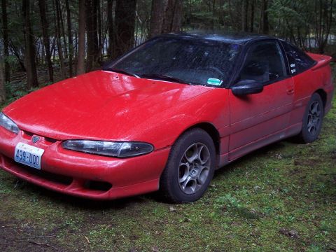 1992 Mitsubishi Eclipse - A red 1992 Mitsubishi Eclipse coupe with a black roof sits in a wooded area, showing its 3/4 front view.