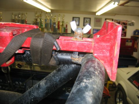 1992 Ford Mustang - Close-up of a red metal bracket secured with a strap and wing nut, with a white car visible in the background.