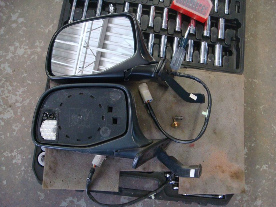 Two car side mirrors, one with glass, are disassembled on a workbench with tools