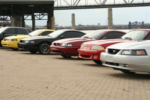 2003 Mercury Marauder - A line of Ford Mustangs are parked on a paved area near a waterfront, with a bridge in the background.