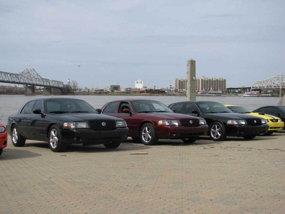 Four Mercury Marauders and a yellow Ford Mustang parked by a river with a bridge in the background