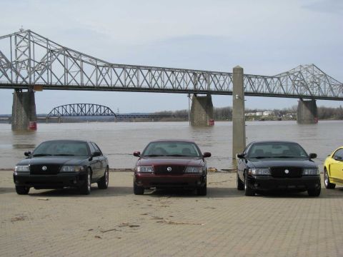 2003 Mercury Marauder - Several Mercury Marauder sedans are lined up by the water with a large bridge in the background.