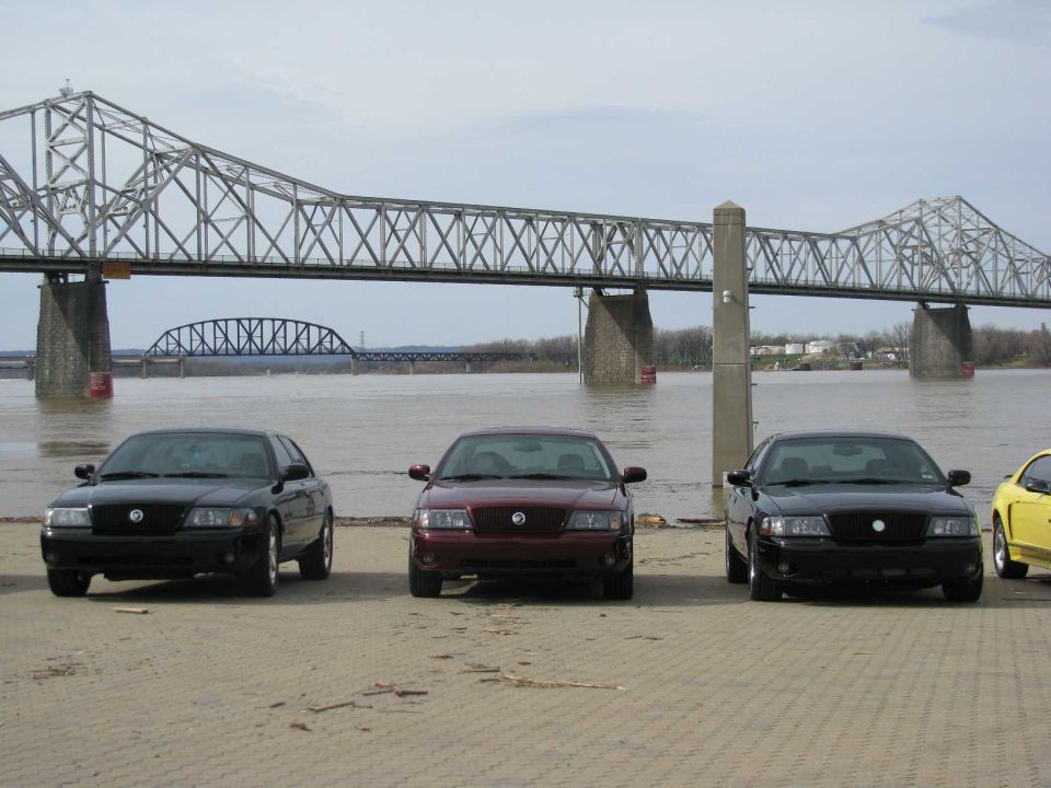 Three black and maroon Mercury Marauders parked in front of a bridge over a river. A yellow Mustang is partially visible on the right