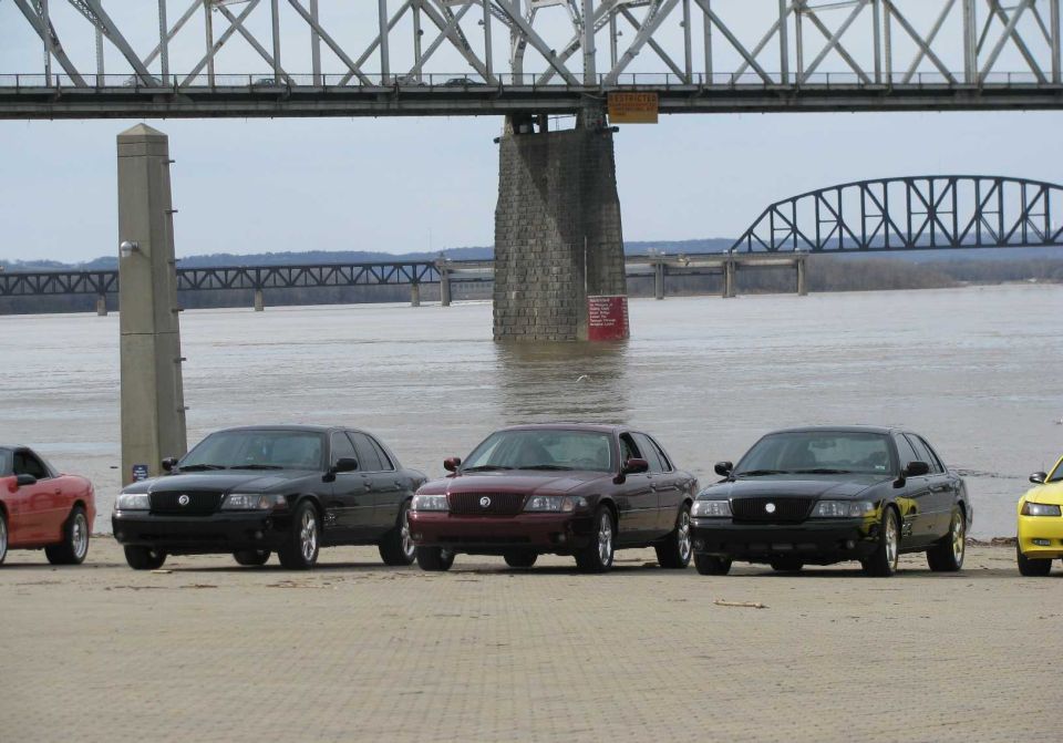 Three black and maroon 2003 Mercury Marauders and a red car parked on a sandy area near a bridge and river