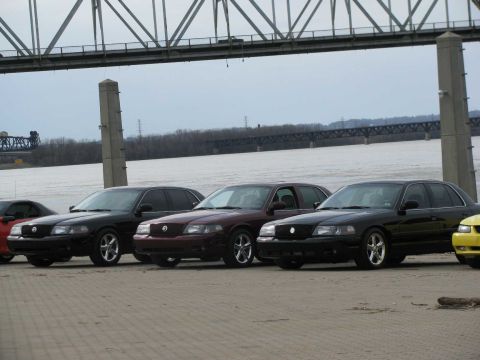 2003 Mercury Marauder - Several Mercury Marauders are parked in a line next to a body of water with a bridge overhead.
