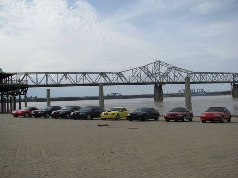 2003 Mercury Marauder - A group of Mustangs are lined up on a paved area near a large bridge over a river.