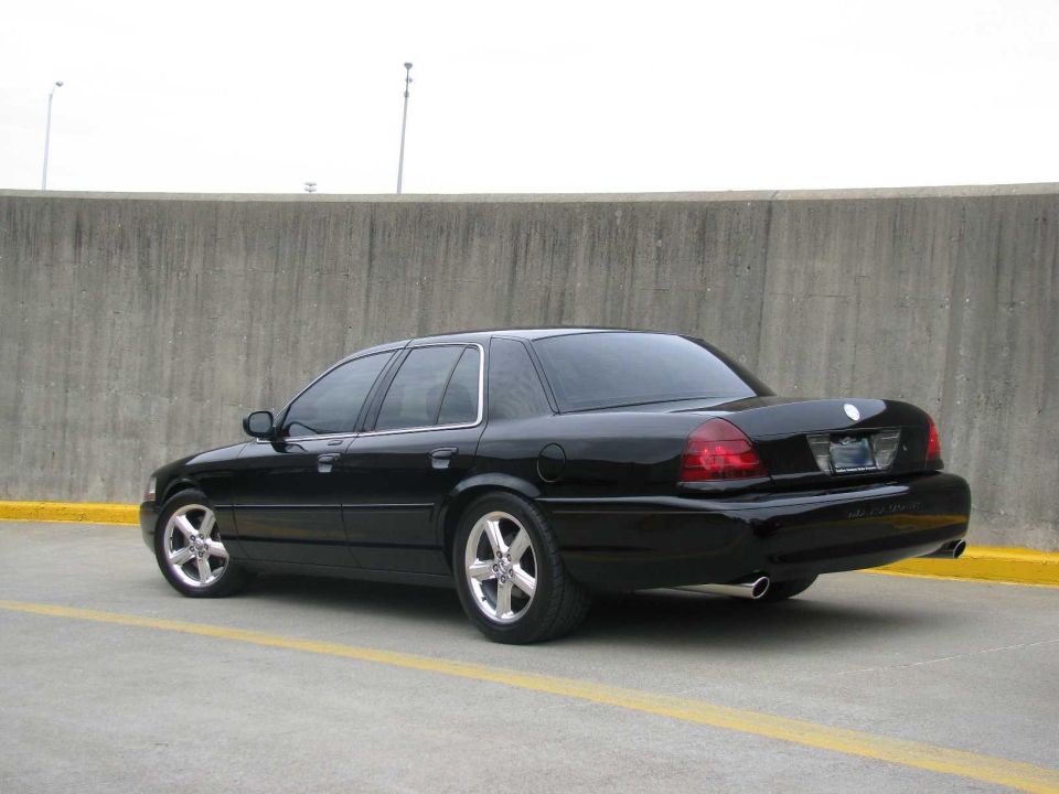 Black sedan parked near a concrete wall. It has aftermarket wheels and dual exhaust pipes