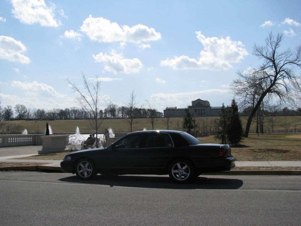 Black sedan parked on the side of the road with a large building and fountains in the background
