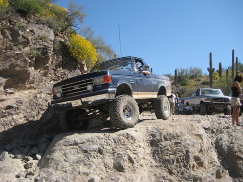 1991 Ford Bronco - Two lifted Ford trucks navigate a rocky desert trail under a clear blue sky.