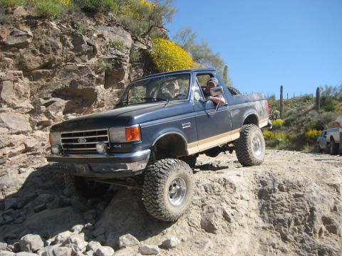 1991 Ford Bronco - A blue Ford Bronco navigates a rocky off-road trail under a clear blue sky.