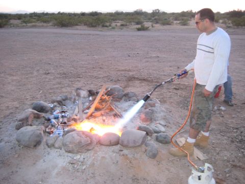1991 Ford Bronco - Man uses a propane torch to start a campfire in a rocky pit, with cans scattered around.
