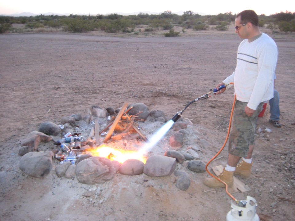 Man with propane torch igniting campfire in desert landscape. Cans and debris surround the fire pit