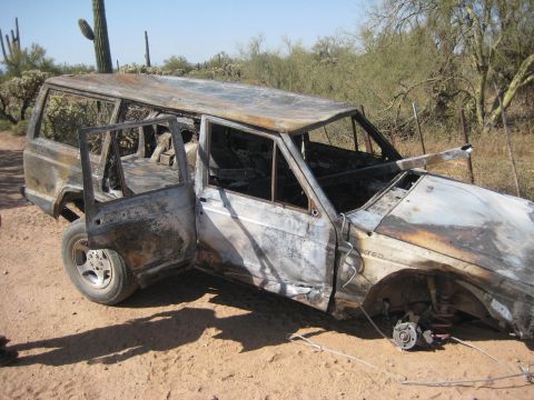 1991 Ford Bronco - A heavily damaged SUV sits in a desert environment, its interior gutted by fire and one wheel missing.