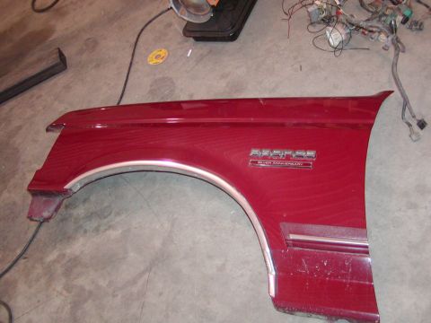 1991 Ford Bronco - A removed burgundy front fender lies outer-side-up on a concrete shop floor, wearing chrome wheel-arch and rocker moldings plus a "Bronco S…