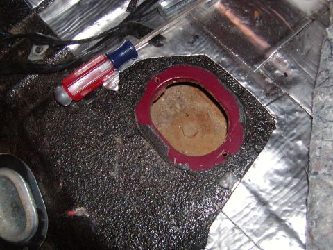 1991 Ford Bronco - Interior teardown shot of a black jute-backed carpet or insulation pad pulled up to expose a red-painted floor opening with a body plug or…