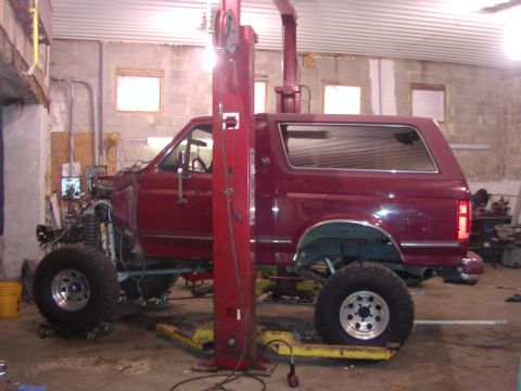 1991 Ford Bronco - A burgundy full-size Ford Bronco sits mid-teardown on a two-post lift in a block-walled shop, driver's side view with the front fender, hoo…