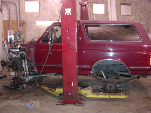 1991 Ford Bronco - Driver's-side view of a burgundy full-size Ford Bronco sitting on a two-post lift with all four wheels off, the front fender removed and th…