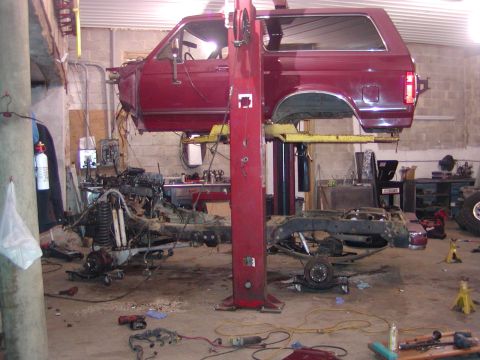 1991 Ford Bronco - A red 1991 Ford Bronco is disassembled on a car lift in a garage, with its engine and chassis removed.