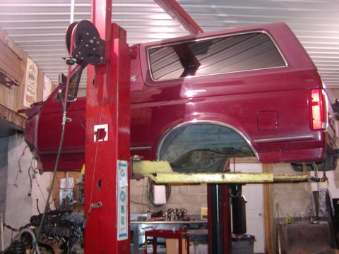 1991 Ford Bronco - A burgundy Ford Bronco body sits high on a red two-post lift with no wheels or drivetrain under it, showing the removable hardtop, chrome w…